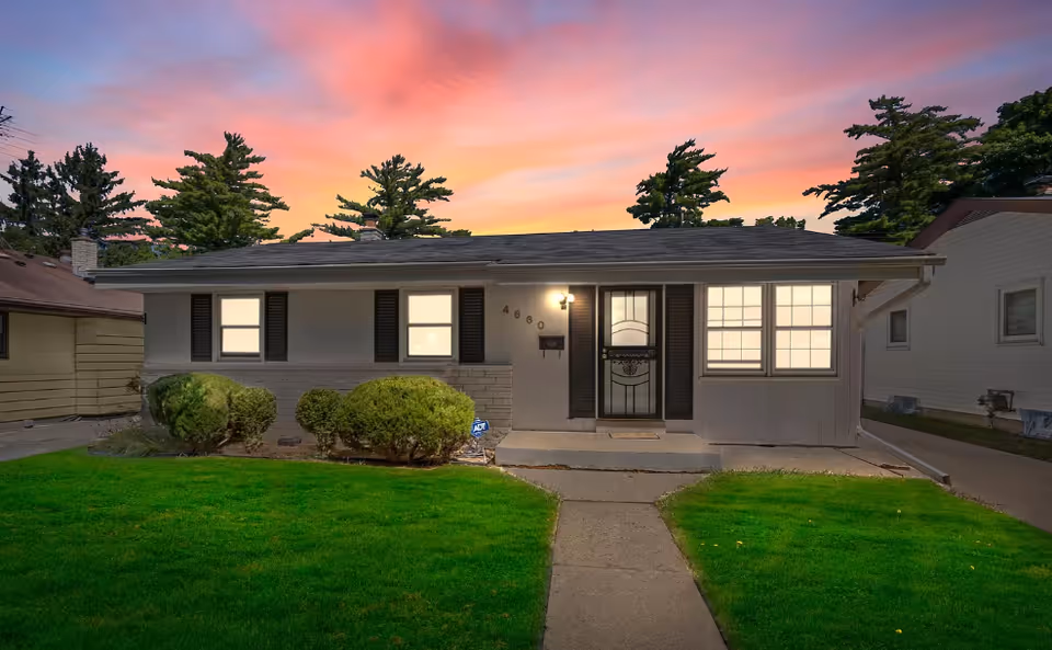 Single-story house with a well-maintained green lawn and bushes in front, illuminated windows, a front door with decorative glass, and a sunset sky with pink and orange hues in the background.