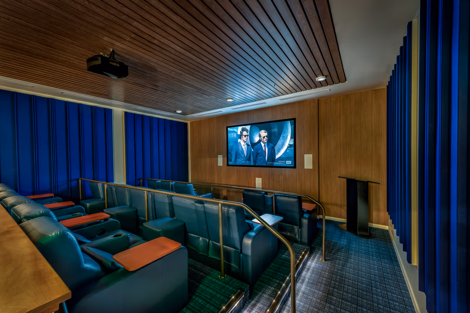 A small, modern movie theater room with blue leather reclining seats arranged in two rows, each seat equipped with a wooden tray. The walls are decorated with blue acoustic panels and wood paneling. A large flat-screen TV is mounted on the front wall, displaying a scene from a movie featuring two men in suits and sunglasses. The ceiling has wooden slats and recessed lighting, and there is a projector mounted above.