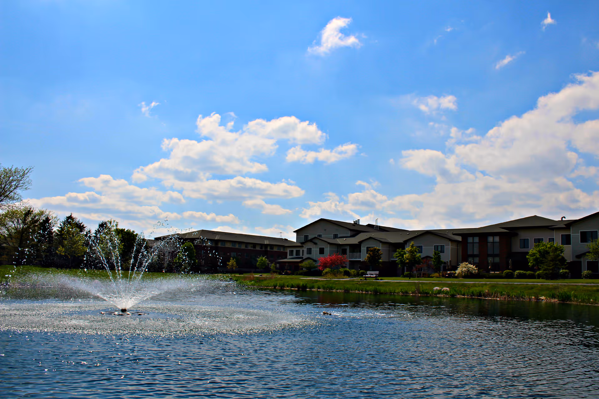 A senior living community building situated behind a pond with a water fountain spraying water into the air. The sky is blue with scattered clouds, and there are trees and shrubs around the pond and building.