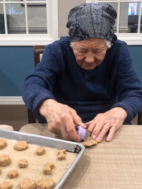 An elderly woman wearing a patterned headscarf and dark sweater is sitting at a table, using a small purple cookie cutter to shape dough on a baking sheet lined with parchment paper. Several shaped dough pieces are visible on the baking sheet in front of her. The background shows a blue and white wall with windows.
