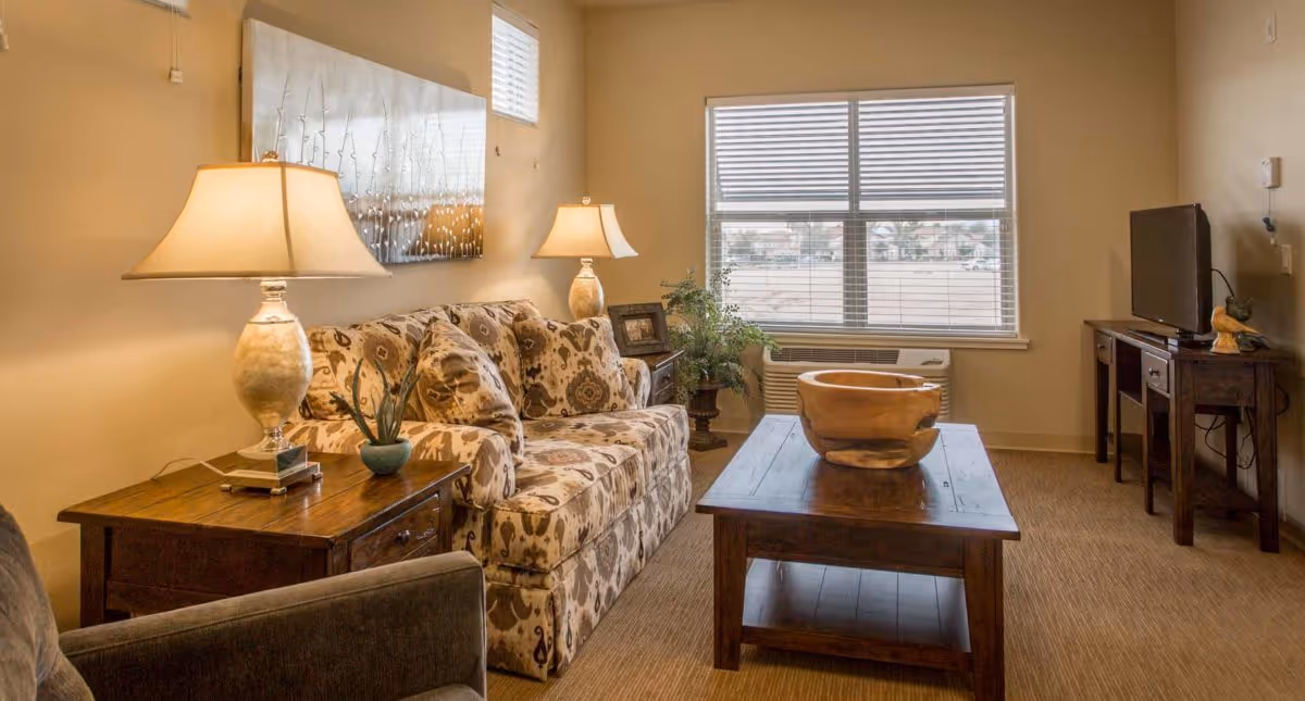A cozy living room with a patterned sofa, two table lamps on wooden side tables, a wooden coffee table with a decorative bowl, a flat-screen TV on a wooden TV stand, a large window with blinds, and a potted plant near the window.