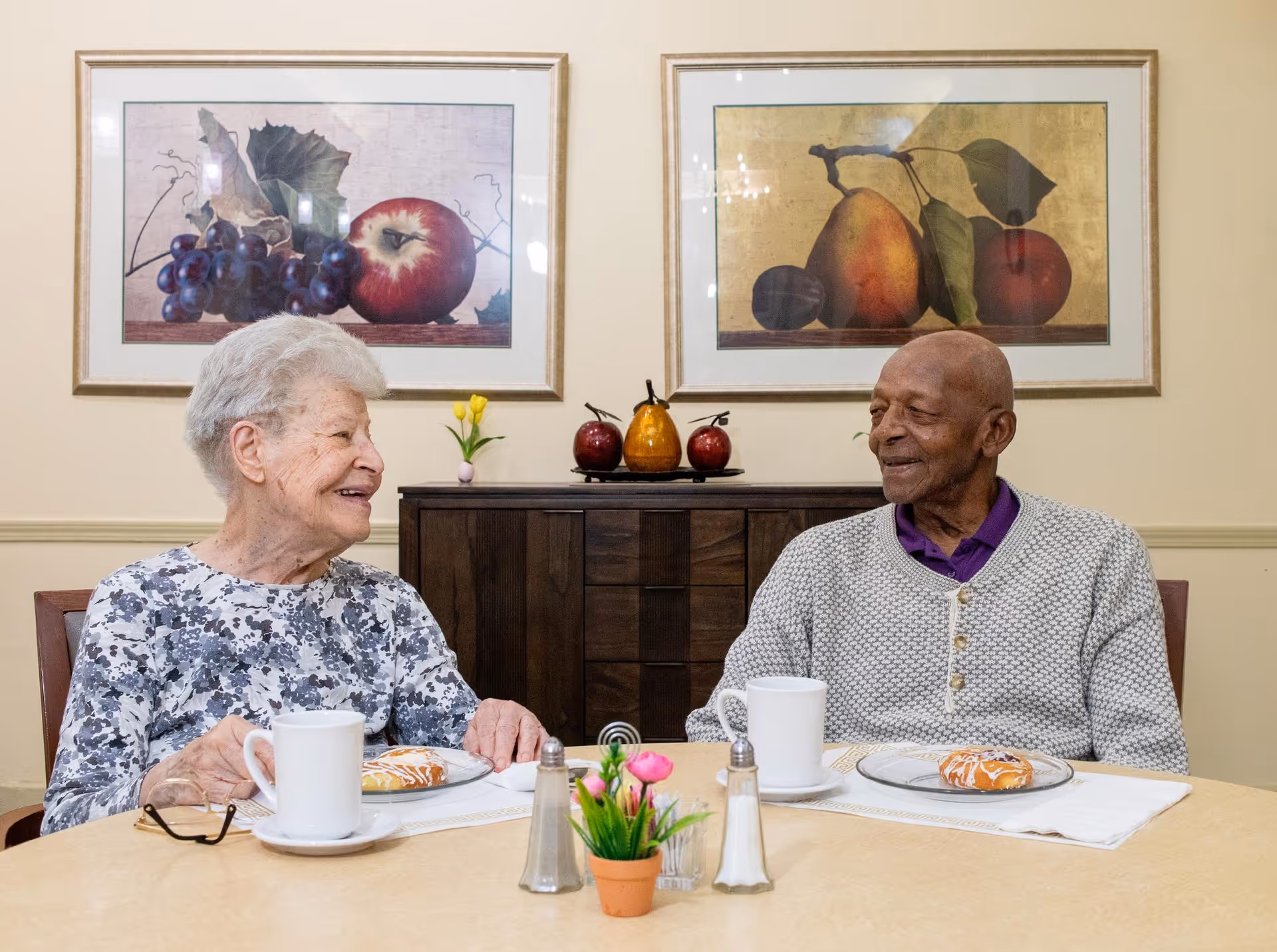Two elderly people sitting at a round table in a dining area, smiling and engaging in conversation. Each has a white mug and a pastry on a plate in front of them. Behind them is a wooden cabinet with decorative fruit and two framed pictures of fruit on the wall.