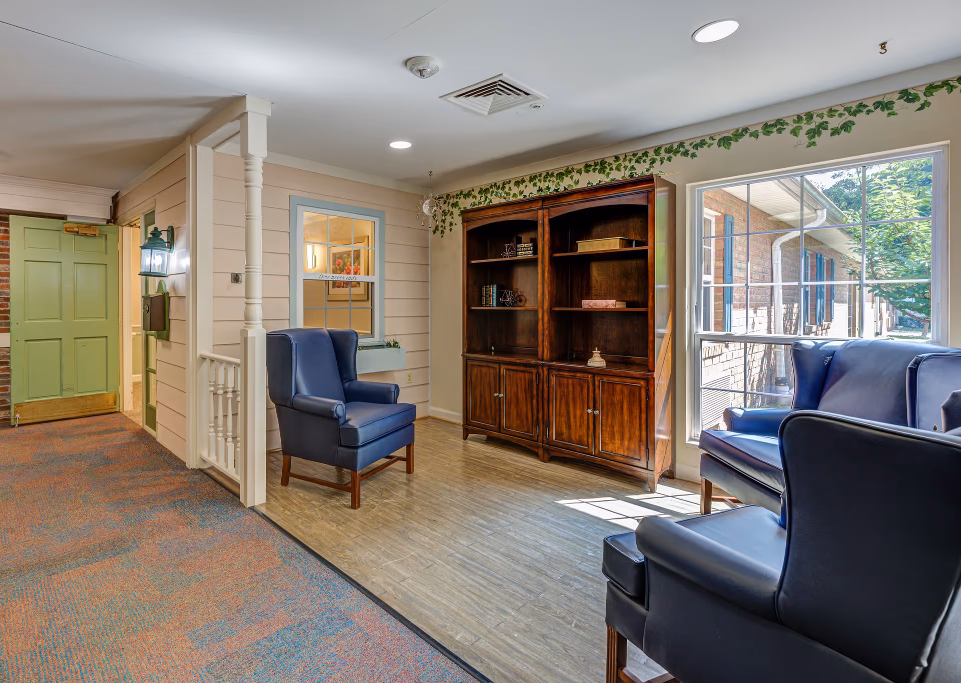 A cozy seating area in a senior living facility with two blue armchairs and a matching ottoman near a large window letting in natural light. There is a wooden bookshelf with decorative items and books, a green door, and a small white railing. The walls are light-colored with a leafy vine border near the ceiling.