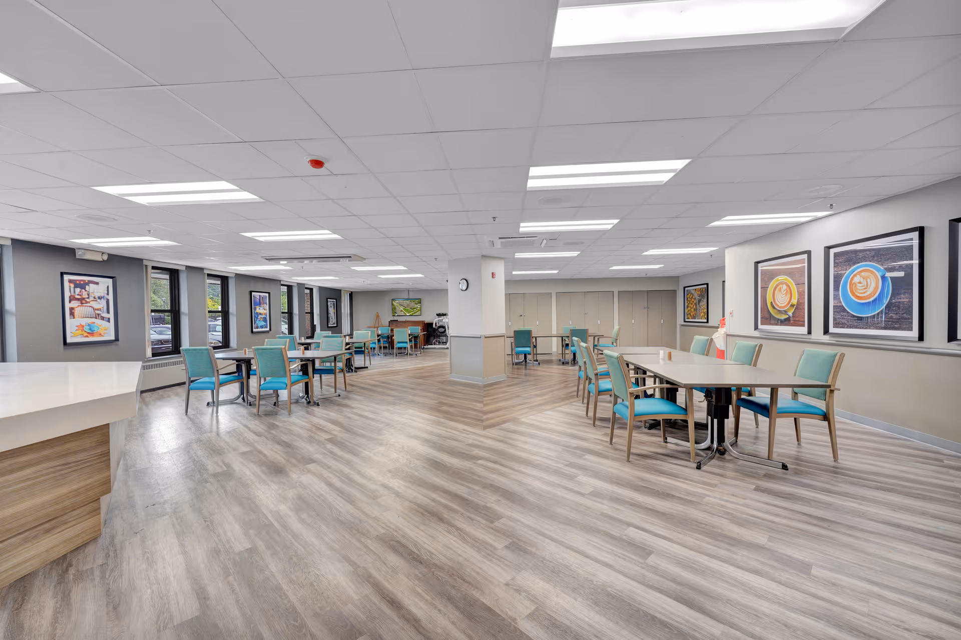 A spacious, well-lit dining area with multiple tables and teal cushioned chairs arranged neatly. The room features large windows along one wall, framed artwork on the walls, and a wood-patterned floor. The ceiling has recessed lighting panels, and there is a white countertop visible on the left side.