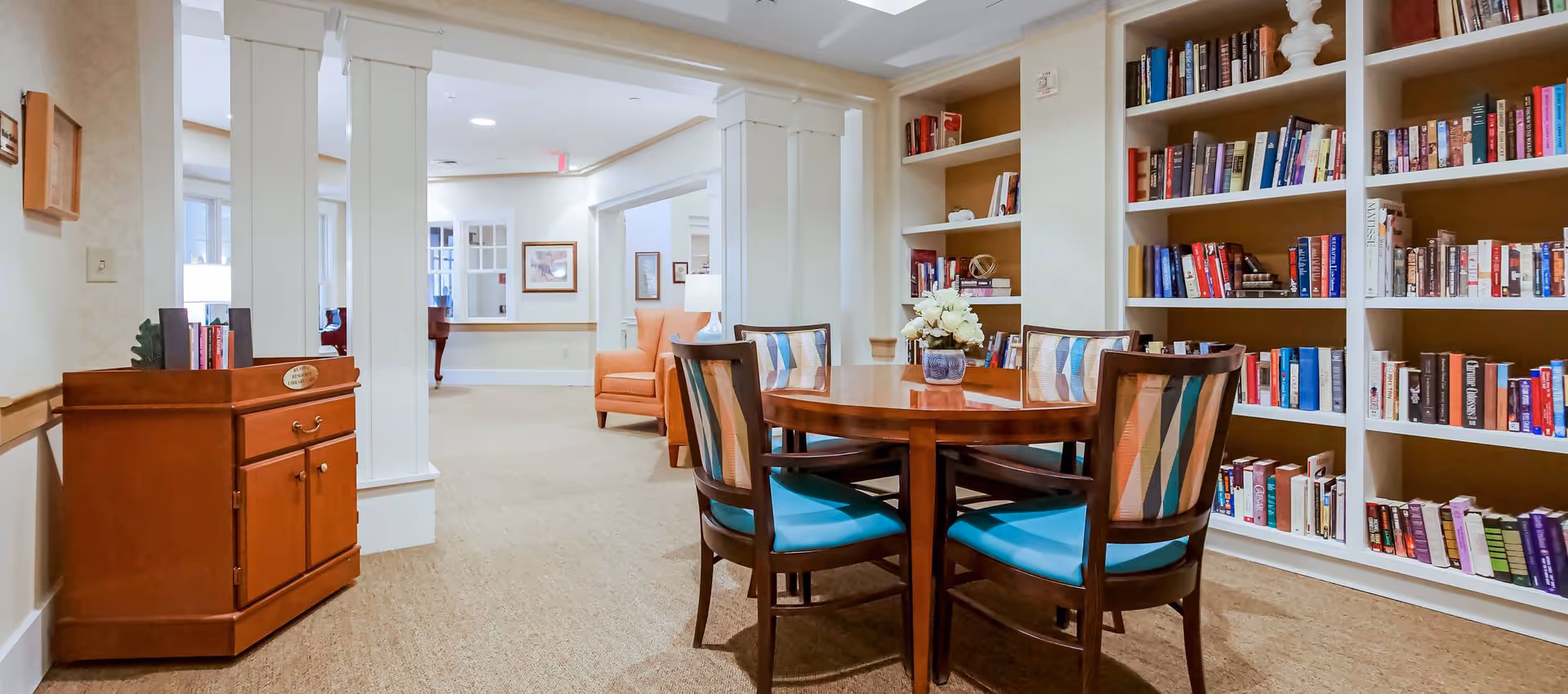 A bright common room featuring a round wooden table with four chairs, built-in bookshelves, and upholstered seating.
