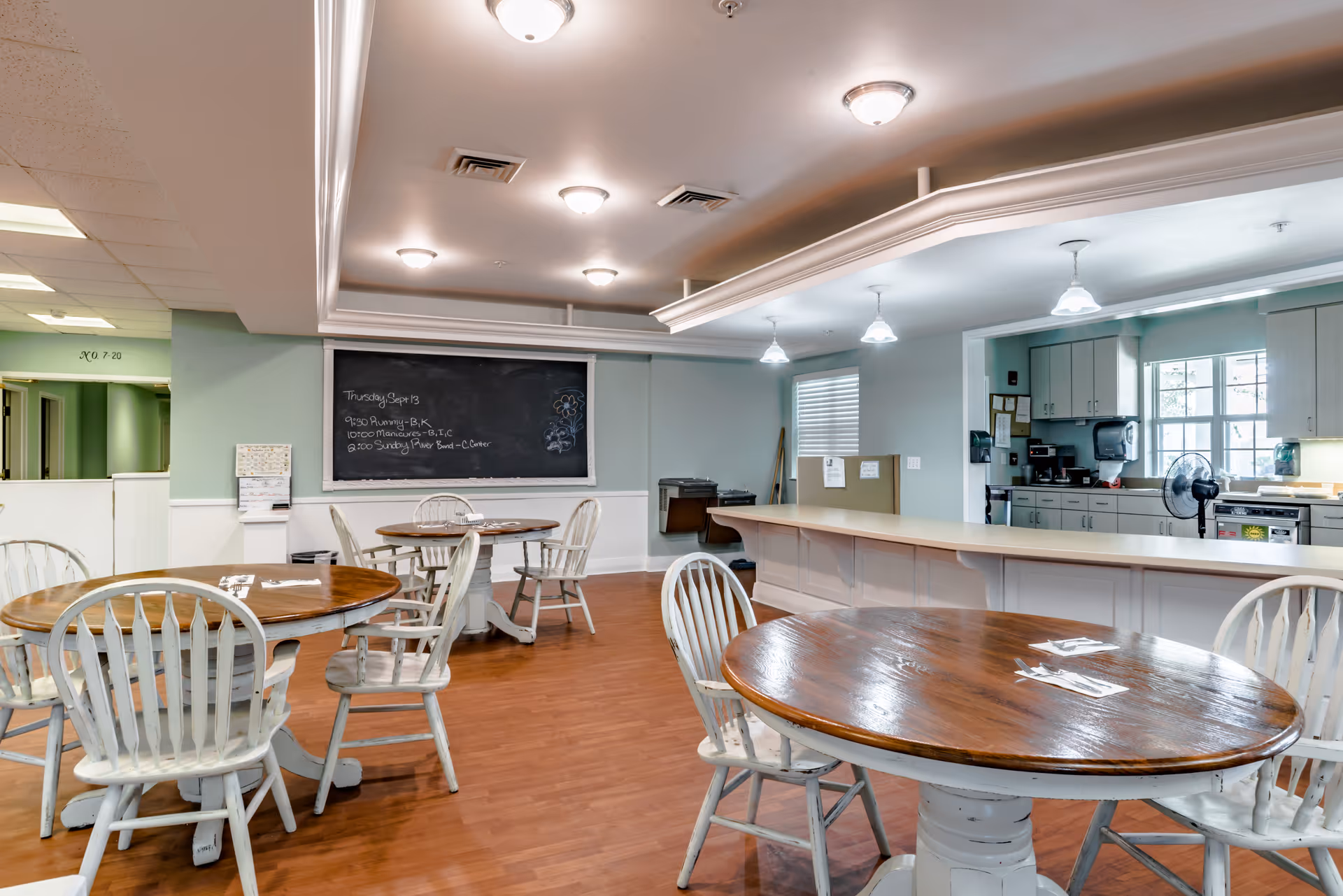 Interior view of a dining area in a senior living facility with round wooden tables and white chairs. The room has a light green wall with a blackboard displaying a schedule, a long counter with pendant lights above, and a kitchen area in the background with cabinets, a window, and appliances.