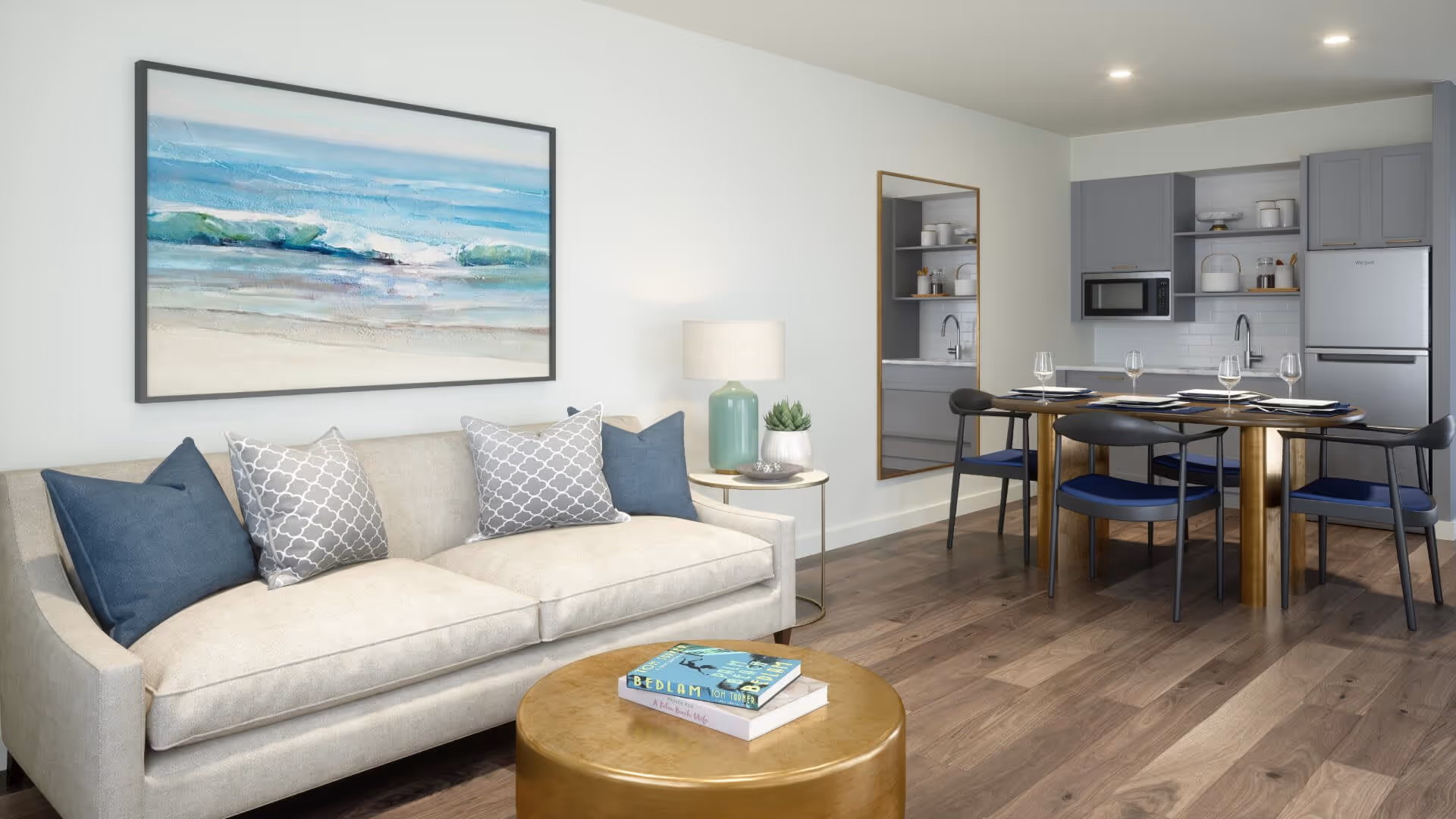 A modern living and dining area featuring a beige sofa with blue and gray patterned pillows, a round gold coffee table with two books on it, a side table with a green lamp and a small plant, a large wall mirror, and a dining table set for four with blue cushioned chairs. The background includes a compact kitchen with gray cabinets, a microwave, and a refrigerator.