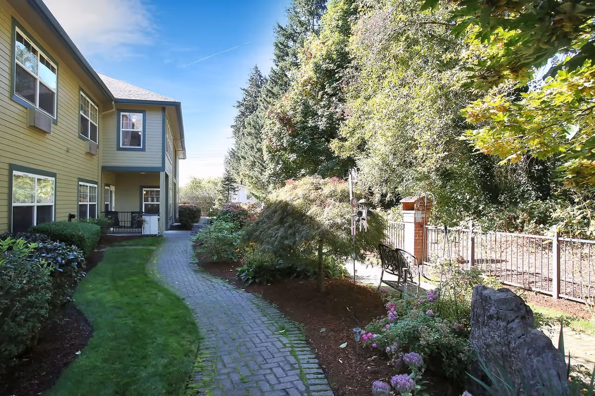 A landscaped outdoor pathway beside a two-story yellow building with green trim. The path is paved with bricks and bordered by green grass, bushes, flowers, and trees. There is a bench and a decorative wind chime near a metal fence on the right side.