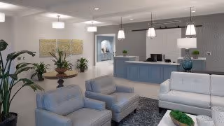A modern and inviting senior living facility common area featuring comfortable seating with two blue armchairs and a white sofa arranged around a coffee table with decorative plants. In the background, there is a reception desk with pendant lights hanging above, and neutral-toned walls with artwork and plants adding to the welcoming atmosphere.