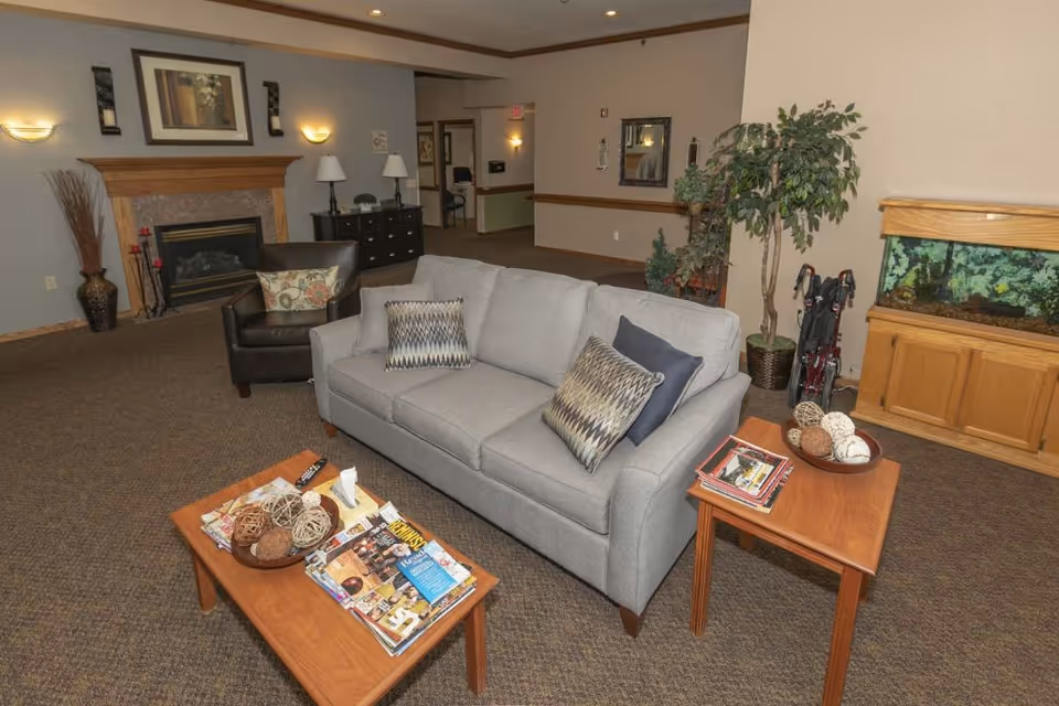 A cozy living room area with a gray sofa adorned with patterned pillows, a dark brown armchair, a wooden coffee table with magazines and decorative balls, and a side table with more magazines and decorative items. The room features a fireplace with a wooden mantle, wall sconces, a large fish tank on a wooden cabinet, and a potted plant. The walls are painted in neutral tones and the floor is carpeted.