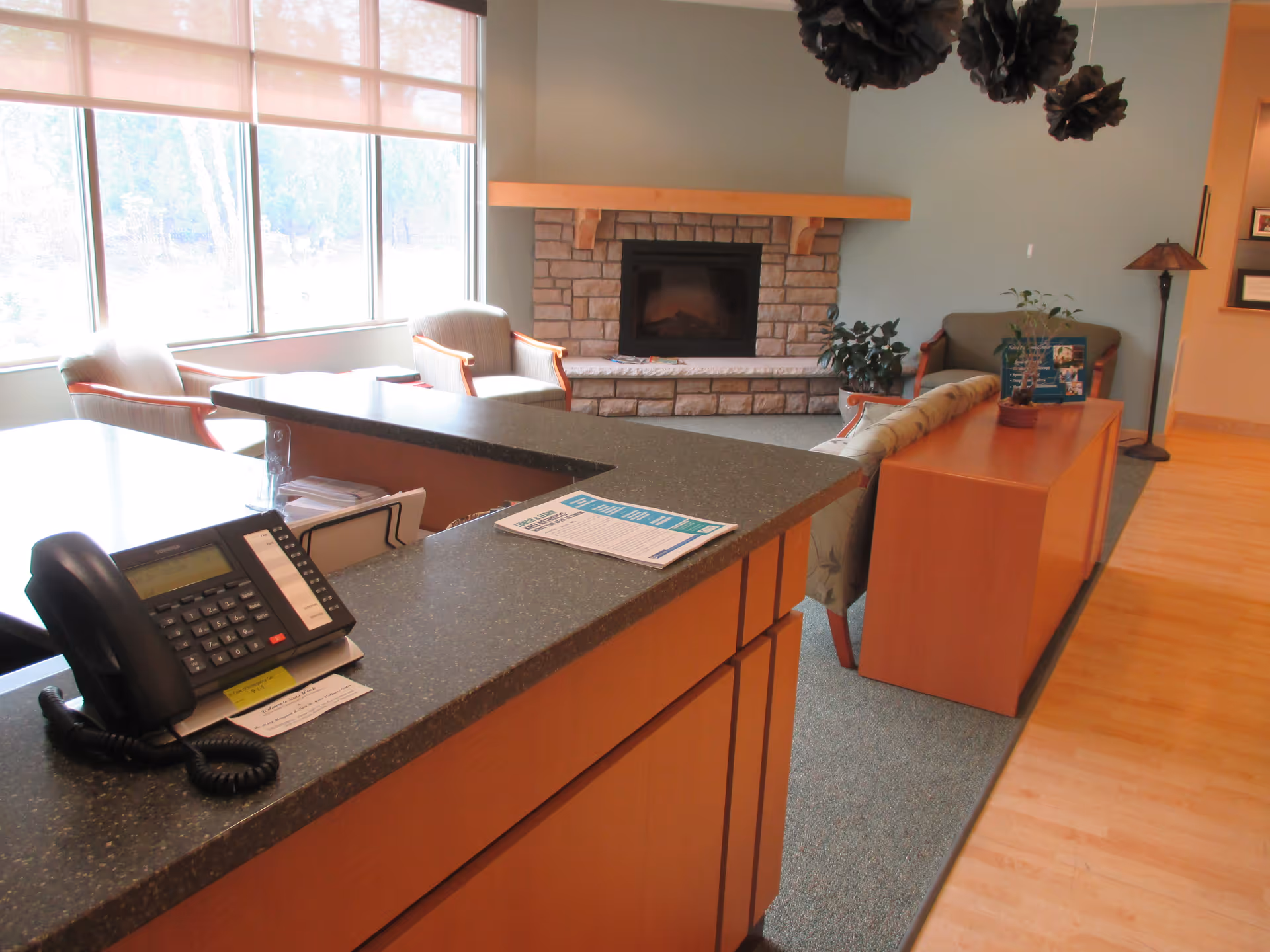 Reception area with a countertop holding a telephone and papers, seating area with chairs and a sofa, a stone fireplace in the background, large windows letting in natural light, and decorative hanging elements from the ceiling.