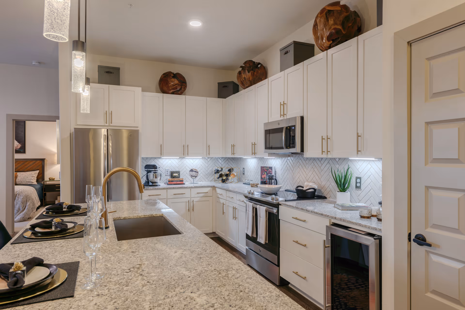 Modern kitchen with white cabinets, granite countertops, stainless steel appliances including a refrigerator, microwave, oven, and wine cooler. The kitchen island has a sink with a gold faucet and is set with plates, napkins, and glasses. Decorative items and a small plant are on the countertops. A bedroom is visible through an open doorway to the left.