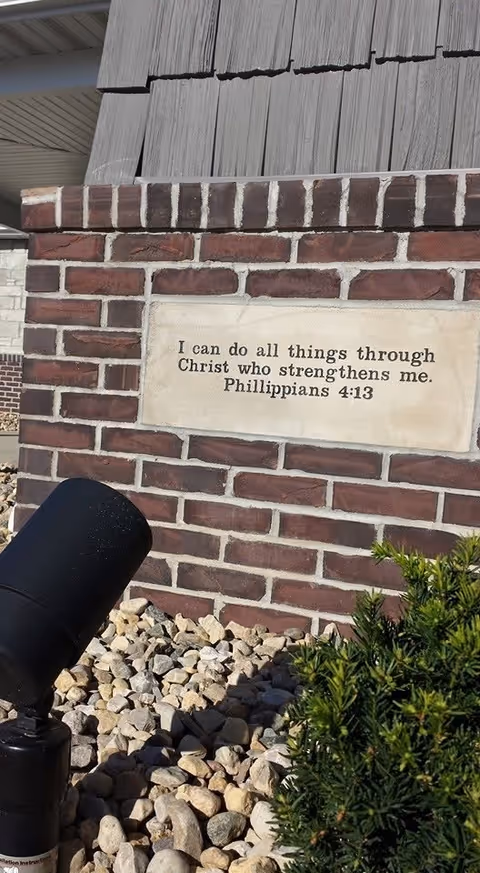 A brick wall with a beige plaque displaying the Bible verse 'I can do all things through Christ who strengthens me. Philippians 4:13'. The wall is surrounded by small rocks and some greenery, with part of a black outdoor light fixture visible in the foreground.