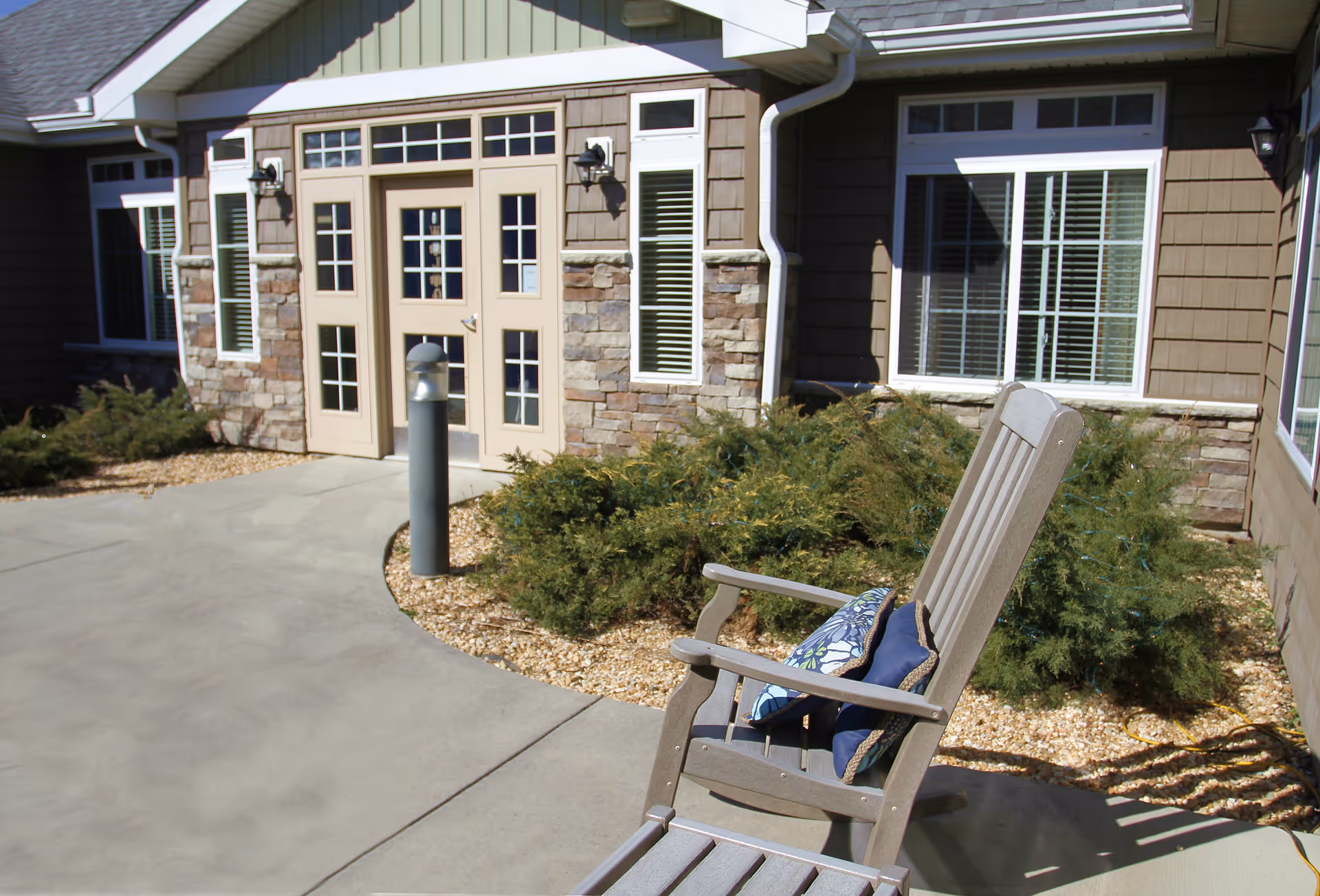 Entrance of a senior living building with a paved walkway, stone-faced facade, windows, and a wooden outdoor chair with cushions.