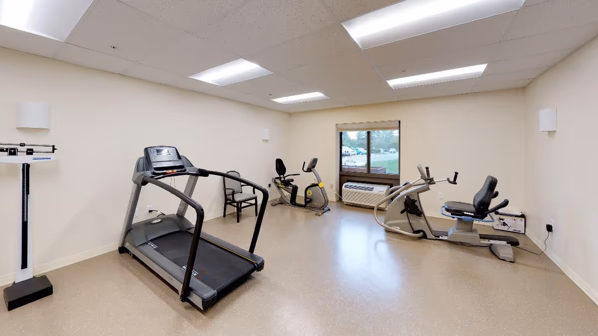A small fitness room with exercise equipment including a treadmill, two recumbent stationary bikes, a weight scale, and a chair. The room has beige walls, a window with a view outside, and ceiling lights.