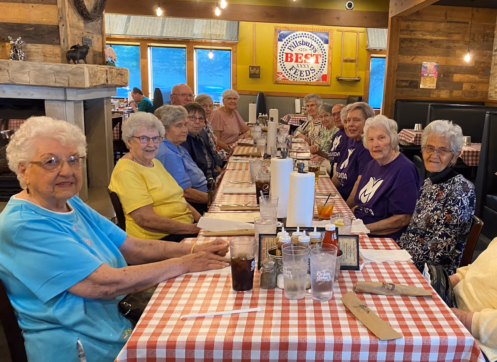 A group of elderly people sitting around a long table with a red and white checkered tablecloth in a restaurant. The table is set with glasses, napkins, condiments, and paper towel rolls. The background shows rustic wooden walls, a fireplace, and a sign that reads 'Pillsbury's Best Feeds'.