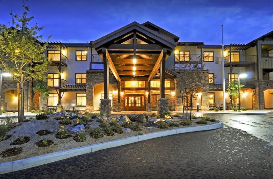Exterior view of Cascades of the Sierra senior living facility at dusk, showing a well-lit entrance with a covered porch supported by wooden beams, surrounded by landscaped garden beds and trees.