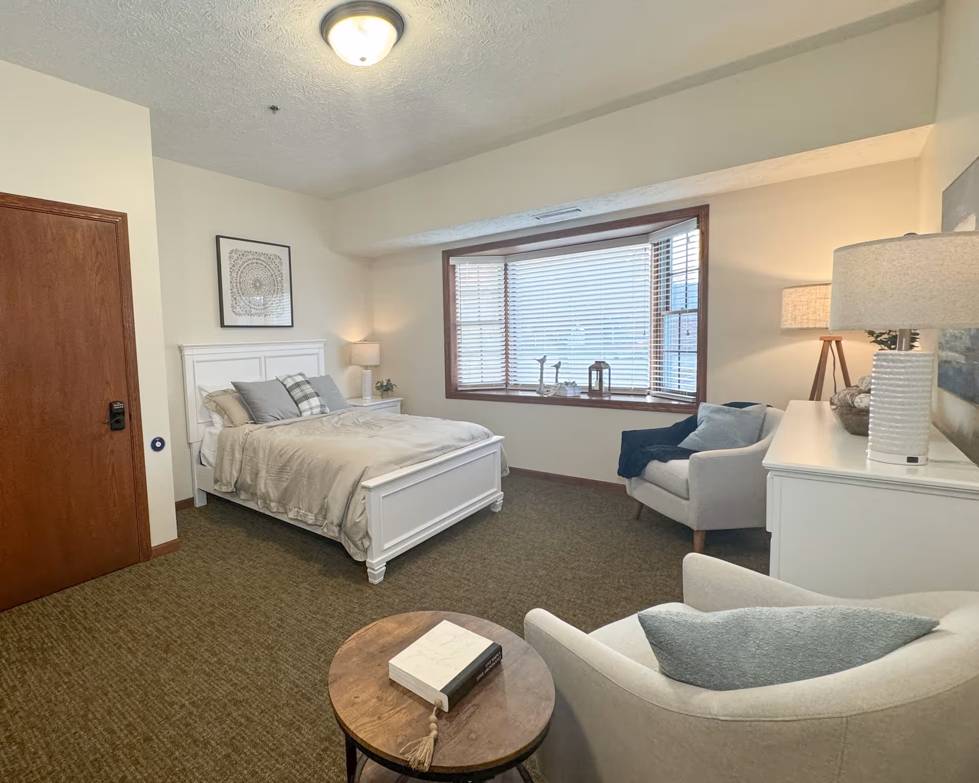 A cozy bedroom with a white wooden bed frame, beige bedding, and multiple pillows. There is a wooden door on the left, a framed artwork above the bed, and a nightstand with a lamp and plant. A large window with wooden blinds lets in natural light. Two upholstered armchairs with cushions are positioned near a round wooden table with a book on it. A white dresser with a lamp and decorative items is on the right side of the room.