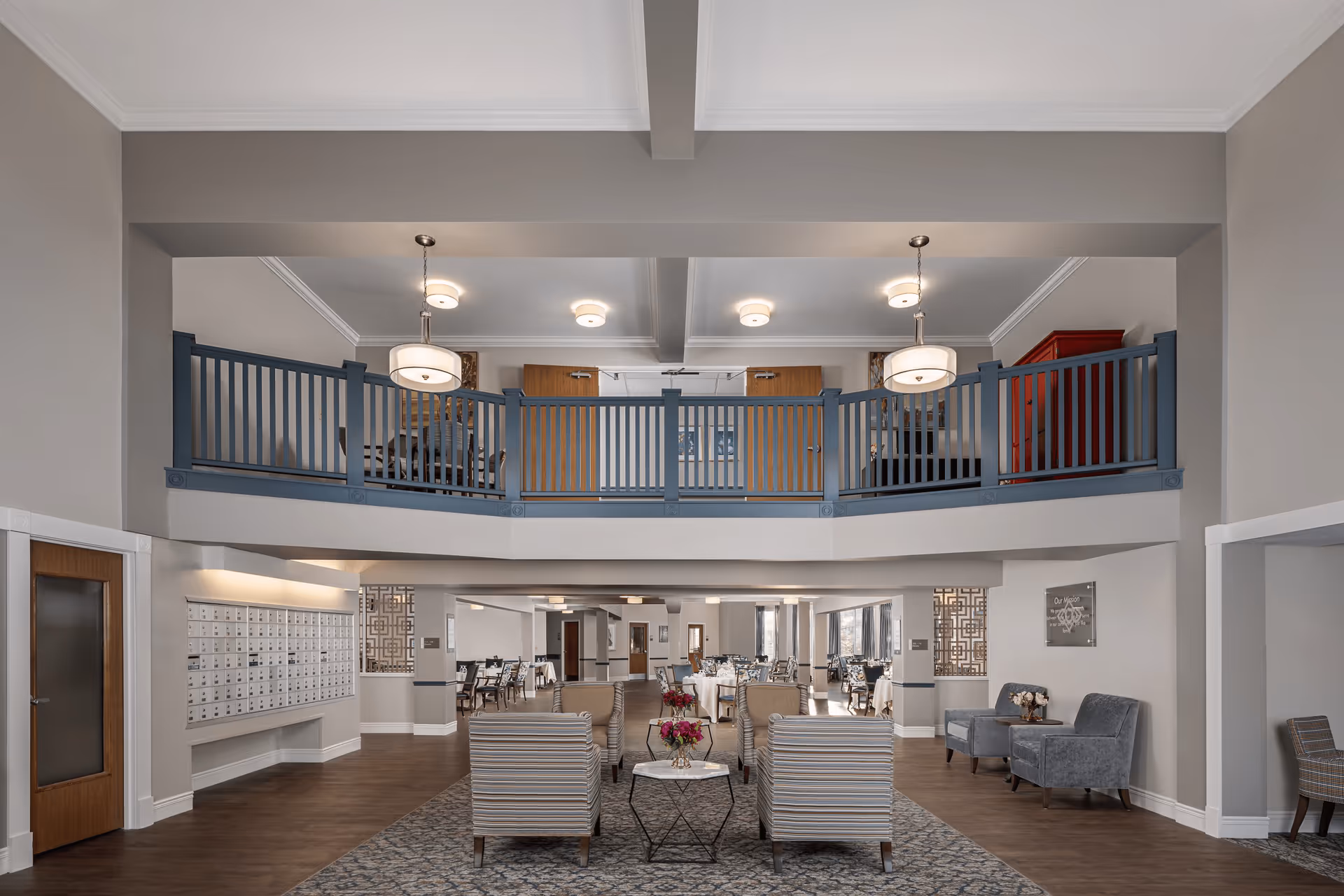 Spacious senior living facility common area with a high ceiling and a mezzanine level featuring blue railings. The lower level has a seating area with striped armchairs and a small table with flowers, a carpeted floor section, and a dining area with tables and chairs in the background. There are mailboxes on the left wall and modern light fixtures hanging from the ceiling.