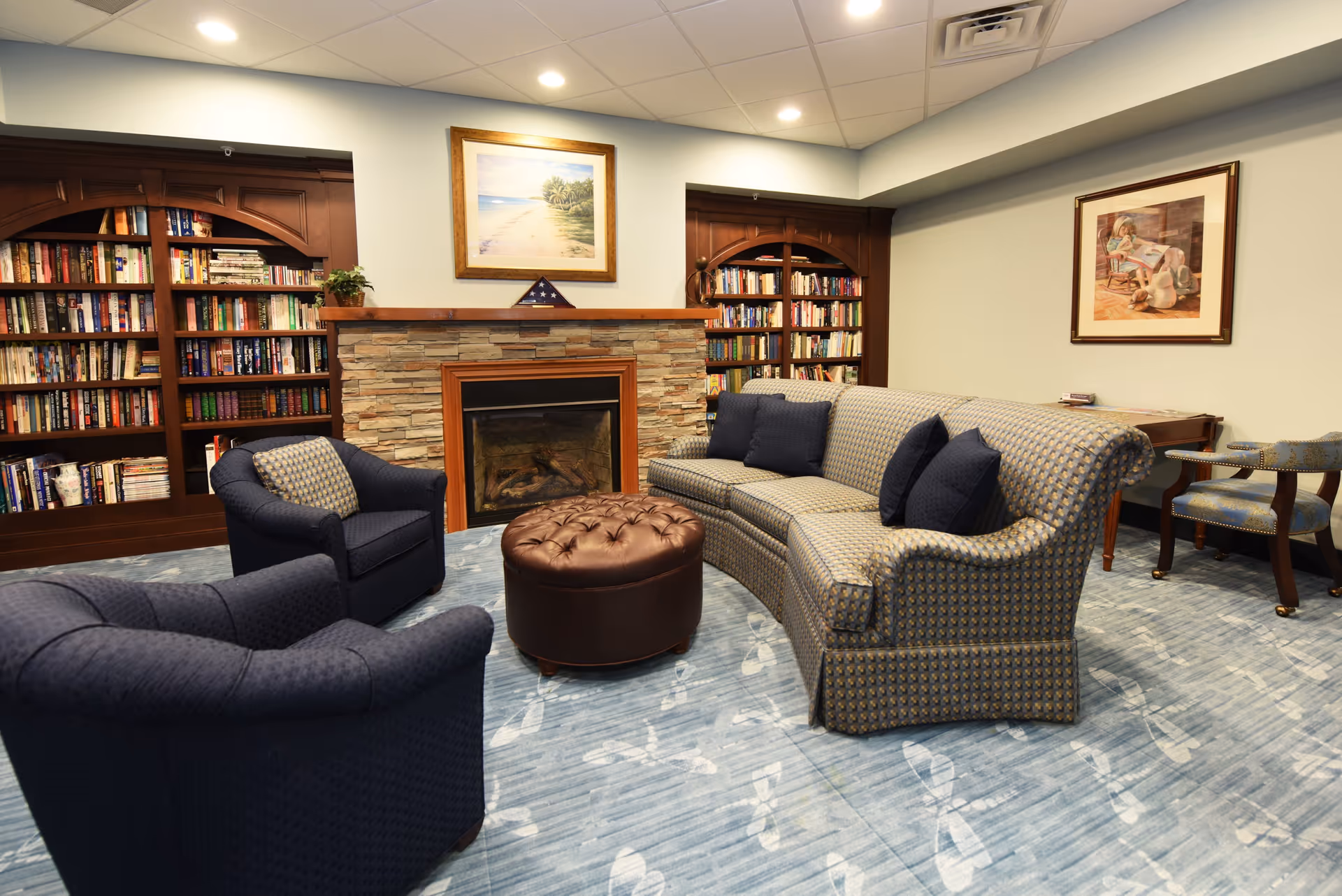 A cozy living room area with a patterned blue sofa, two matching armchairs, and a round brown leather ottoman in the center. Behind the seating is a stone fireplace with a wooden mantle, flanked by built-in wooden bookshelves filled with books. The walls are light-colored with framed artwork hanging above the fireplace and on the adjacent wall. The floor is covered with a blue carpet featuring a subtle leaf pattern.