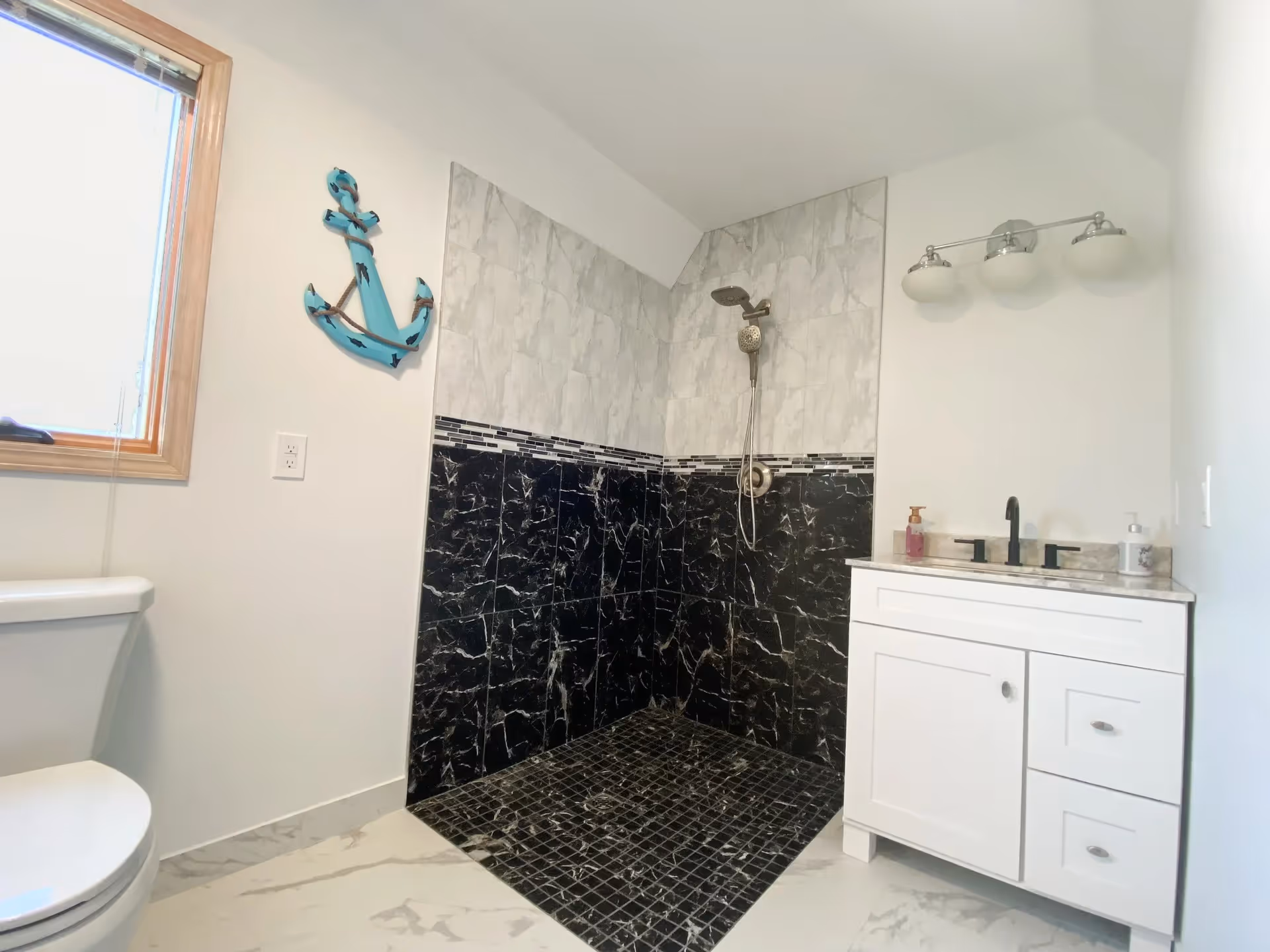 A modern bathroom featuring a walk-in shower with black and white marble tiles, a white vanity with a black faucet, a toilet, a window with a wooden frame, and a blue anchor wall decoration.