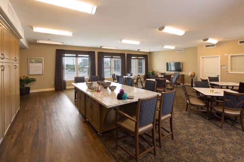 A spacious common area in a senior living facility featuring a large central island with chairs around it, multiple tables with chairs, a flat-screen TV mounted on the wall, large windows with dark curtains allowing natural light, and a mix of wood and carpet flooring.