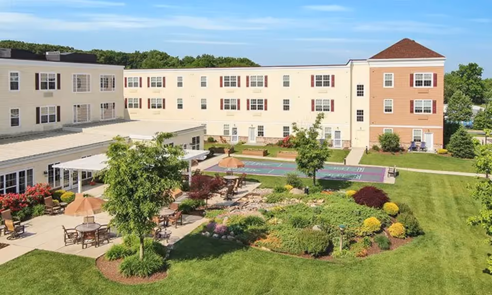 Outdoor courtyard area of a senior living facility with patio tables and chairs under umbrellas, a landscaped garden with various shrubs and flowers, a shuffleboard court, and a three-story building with multiple windows and red shutters in the background under a clear blue sky.