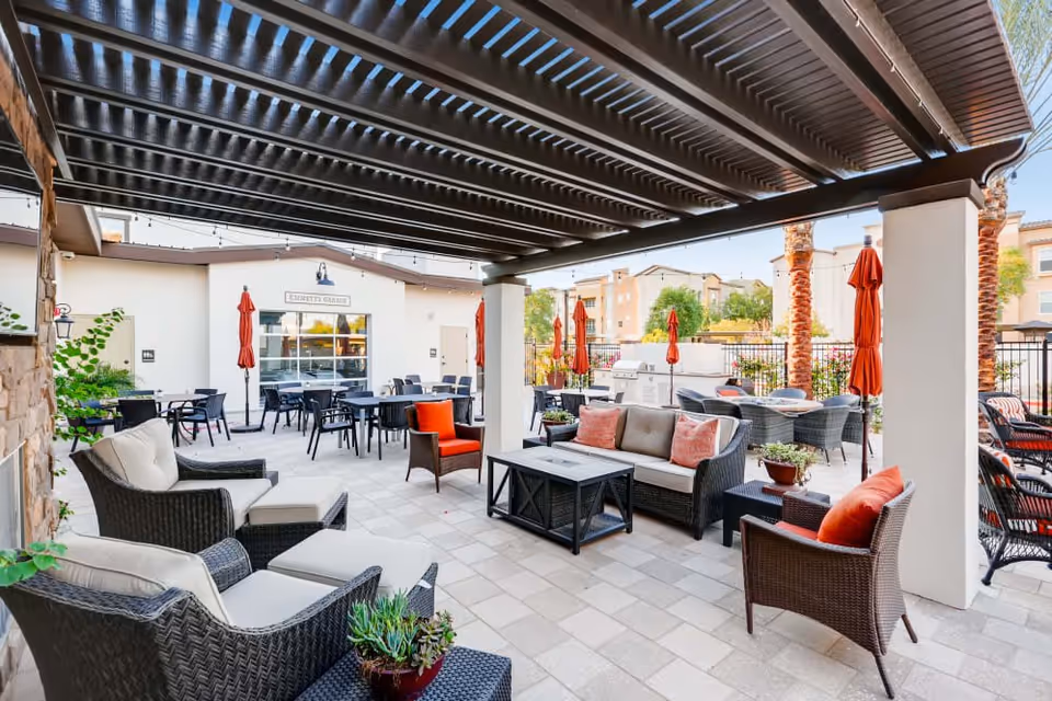 Outdoor patio area with wicker seating including cushioned chairs and sofas under a pergola. Several tables and chairs with red umbrellas are arranged around the space. There are palm trees and a building labeled 'Emmett's Garage' in the background.