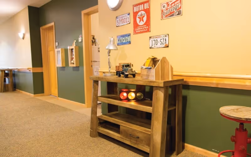 A hallway in an assisted living facility with green and yellow walls, a wooden console table holding a desk lamp, a toy truck, and a decorative traffic light. Vintage license plates and signs are mounted on the yellow wall above the table. A red stool is partially visible on the right side.