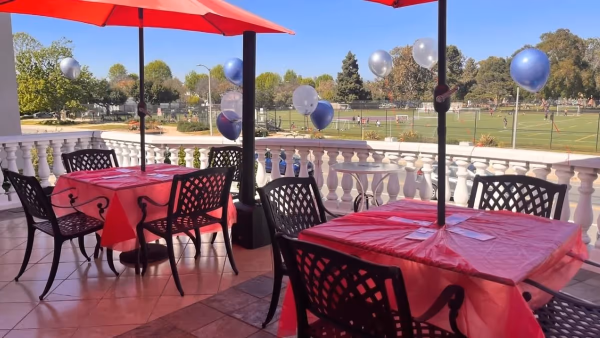 Outdoor patio area with tables covered in red tablecloths and black metal chairs. Red umbrellas provide shade over the tables. Blue and silver balloons are tied to the patio railing. In the background, there is a sports field with people playing and trees under a clear blue sky.