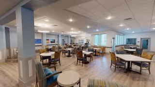 Spacious dining room with multiple tables and chairs arranged neatly. The room has light-colored wooden flooring, soft blue walls, and large windows allowing natural light to fill the space. Ceiling lights provide additional illumination, and there are some comfortable armchairs in the foreground.