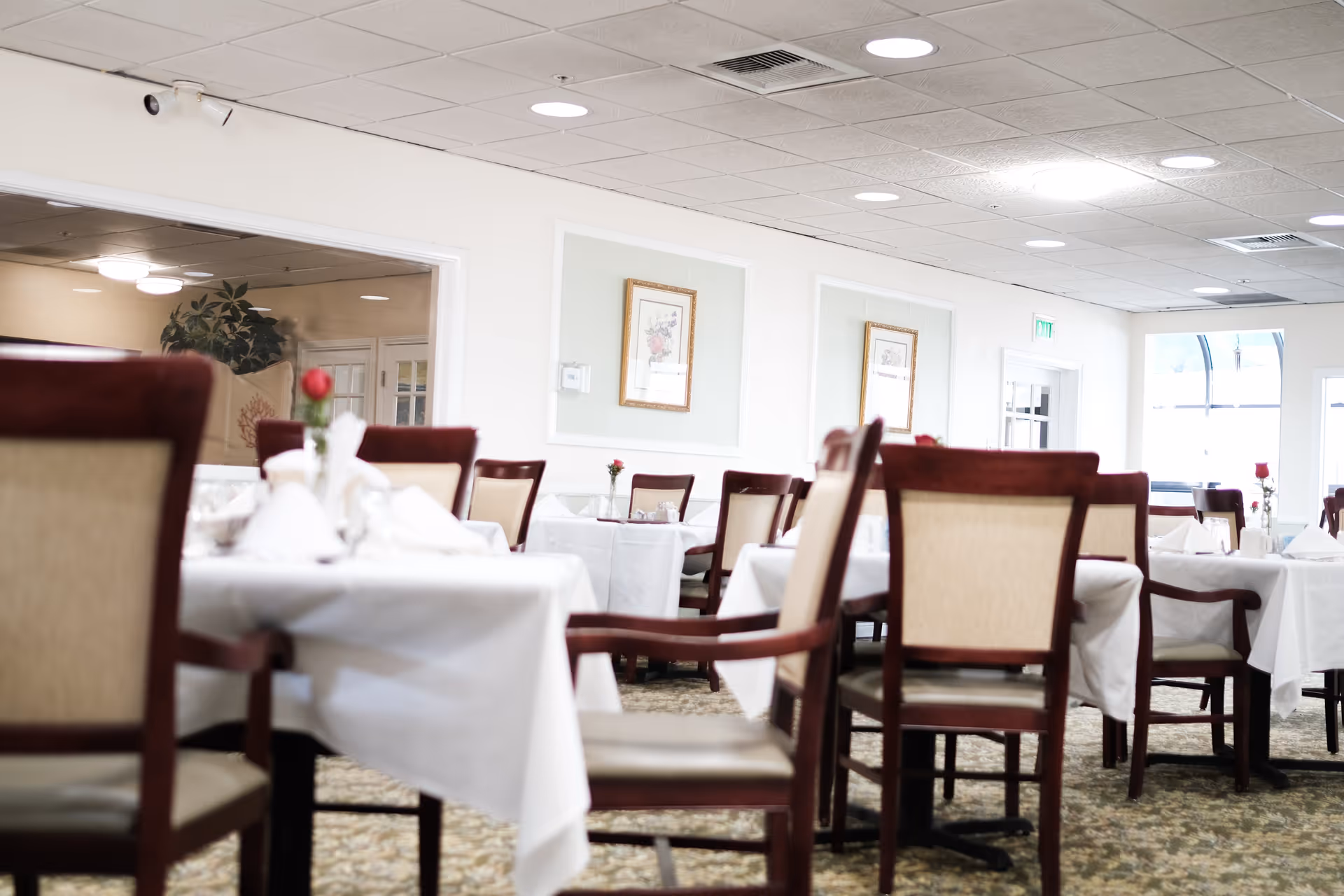 A bright dining room with tables covered in white tablecloths and set with napkins and small vases holding single red roses. The room has wooden chairs with beige cushions, framed artwork on the walls, and a carpeted floor.