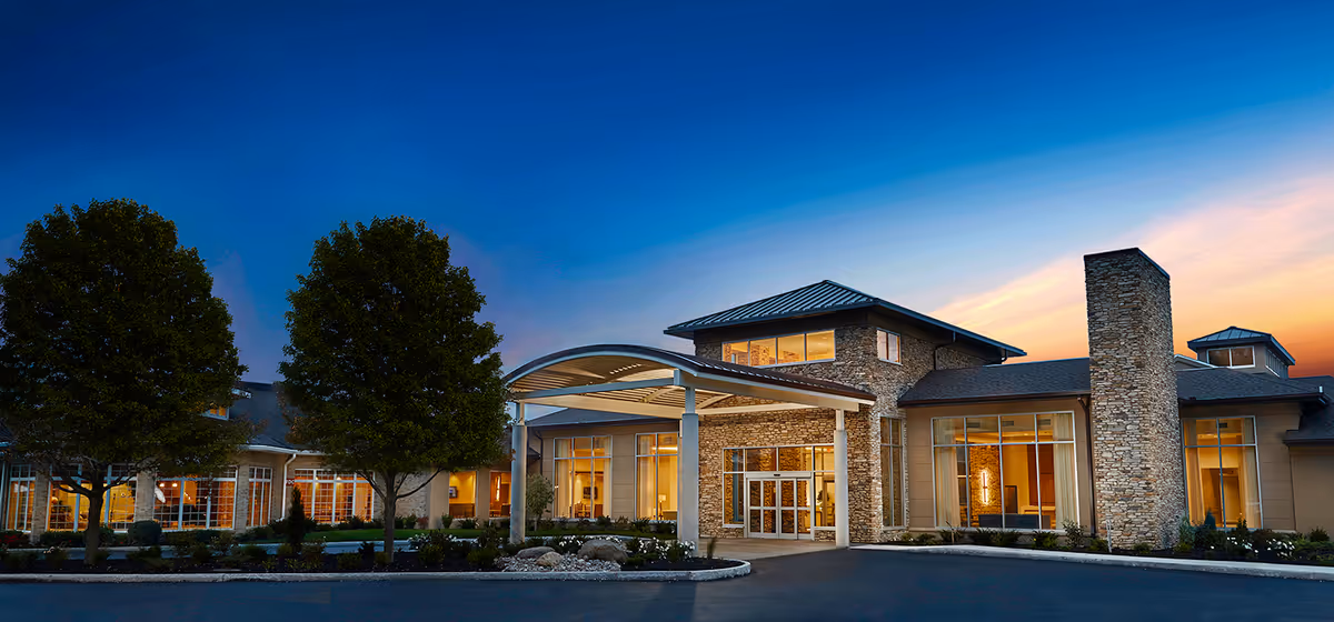 Front entrance of a large brick-and-stone senior living facility at dusk with a covered portico, illuminated windows, and landscaped trees.