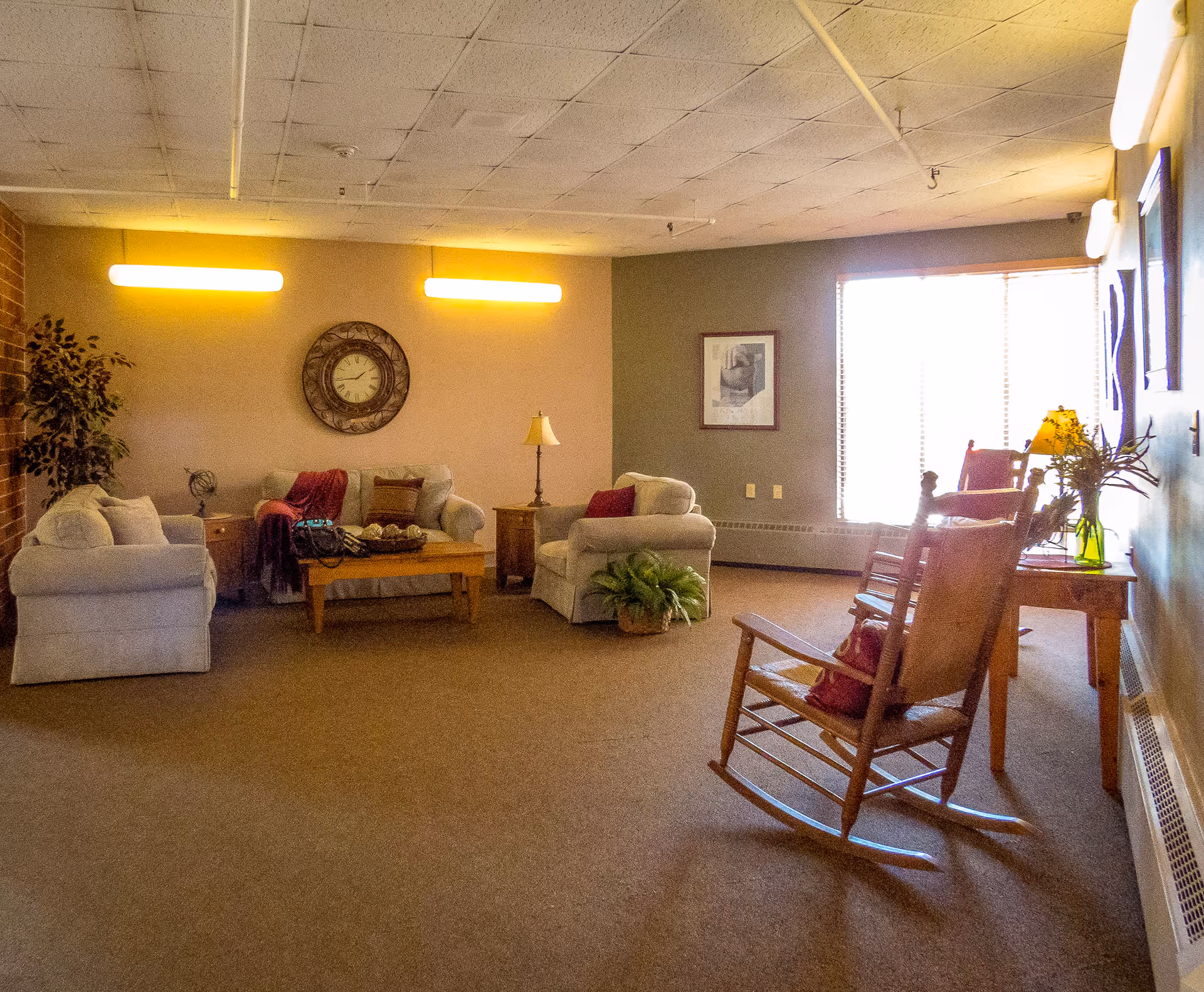 A cozy living room with beige carpet and walls painted in two tones. The room features two beige sofas with cushions and throws, a wooden coffee table, a side table with a lamp, a large wall clock, a potted plant, a wooden rocking chair with a cushion, and a wooden table with a lamp and a vase of flowers near a large window with blinds.