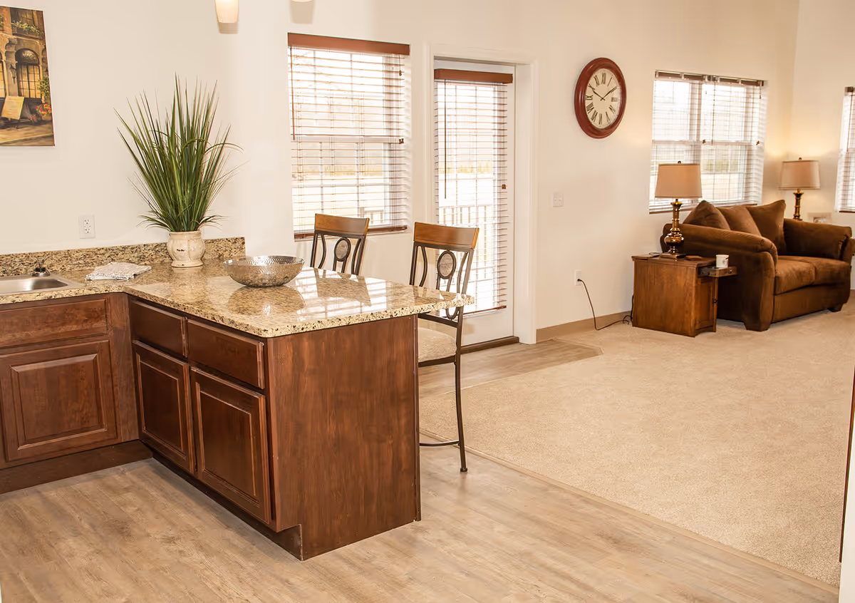 Open-plan living area with a granite-topped kitchen island and bar stools in the foreground and a carpeted seating area with a sofa and side table in the background.