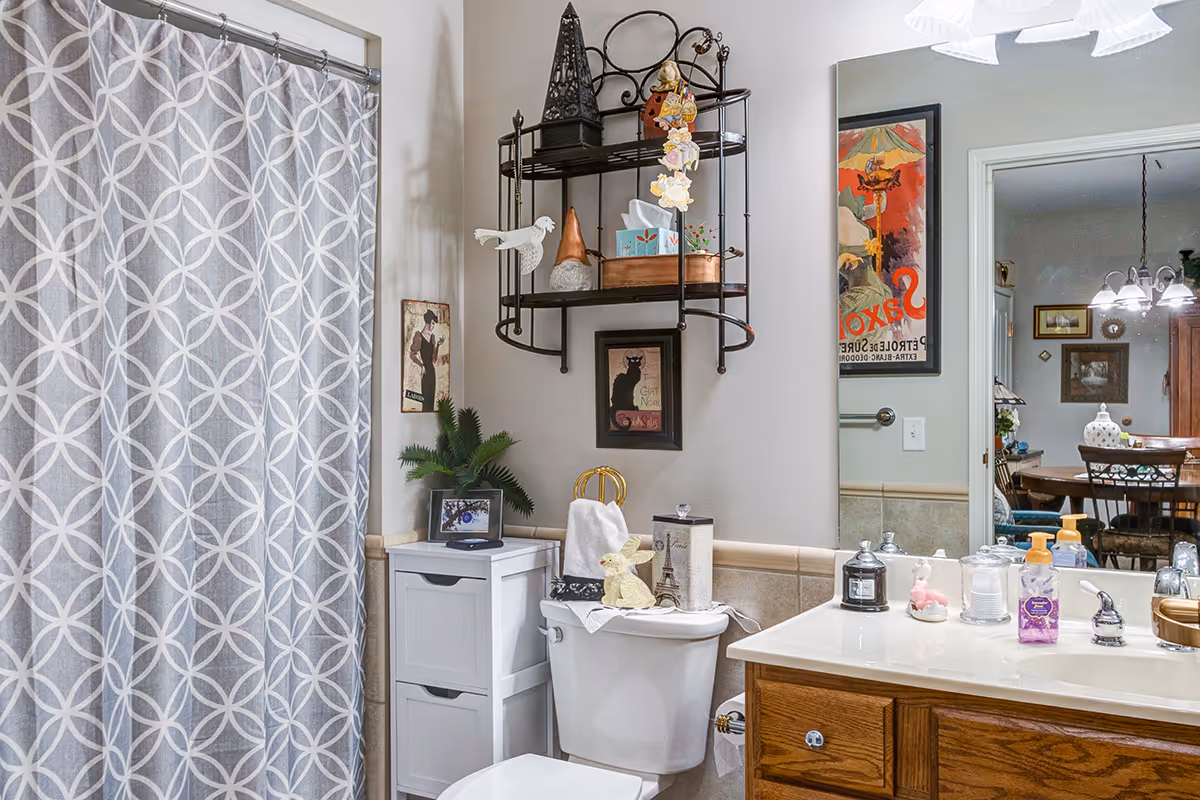 A bathroom with a white toilet, a white storage cabinet, and a wooden vanity with a sink. The shower has a gray and white patterned curtain. Above the toilet is a black metal wall shelf holding decorative items and tissues. The walls have framed artwork, and a large mirror reflects a dining area with a wooden table and chairs.