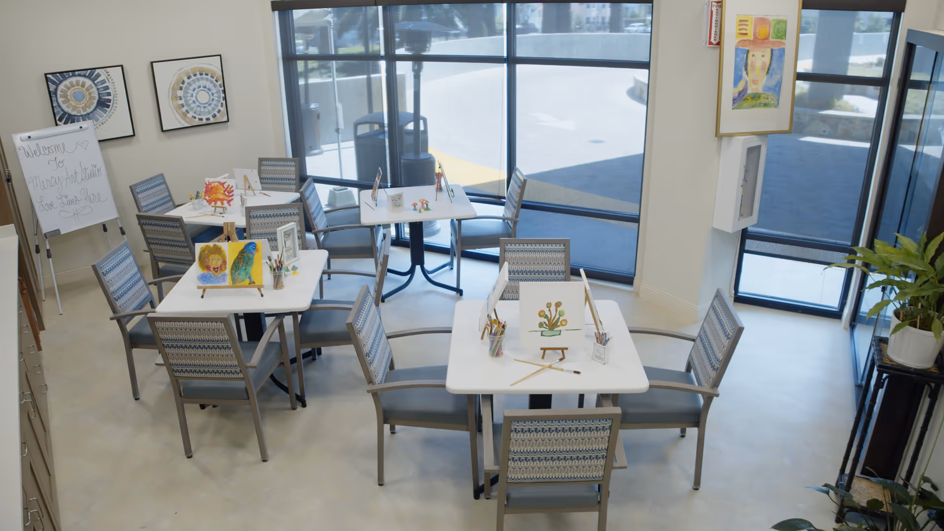 A bright art studio room with several tables and chairs arranged for painting activities. Each table has small easels displaying colorful paintings, paintbrushes, and containers holding art supplies. Large windows allow natural light to fill the room, and there is a whiteboard with a welcoming message. The room has light-colored walls and floor, with framed artwork on the walls and a potted plant near the entrance.
