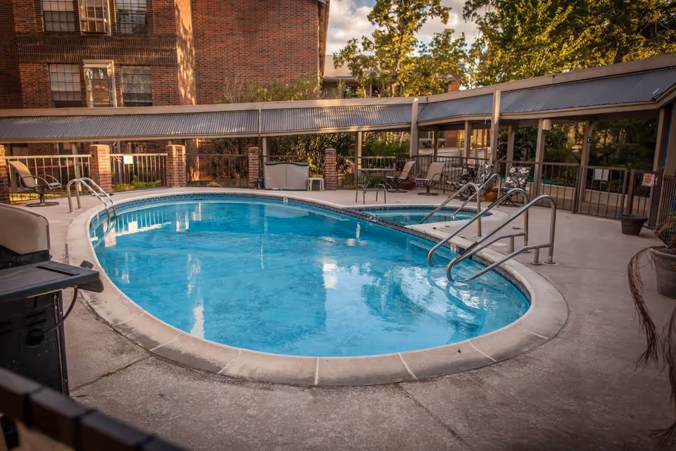 Outdoor swimming pool area with clear blue water, surrounded by a concrete deck. There are metal handrails for pool entry, several chairs, and tables under a covered patio. In the background, there is a brick building and trees with green foliage.