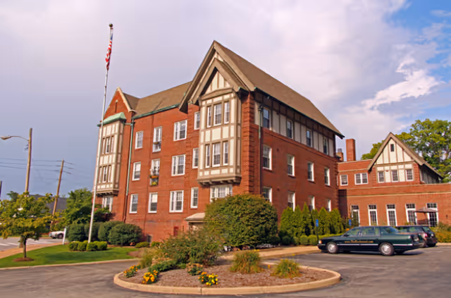 Exterior view of a large brick building with Tudor-style architectural elements, multiple windows, and a steeply pitched roof. There is a flagpole with an American flag in front, surrounded by landscaped greenery and a circular driveway with parked cars.