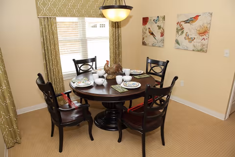 Small dining room with a round wooden table set for four, dark chairs, green curtains at a window, and bird-themed wall art.