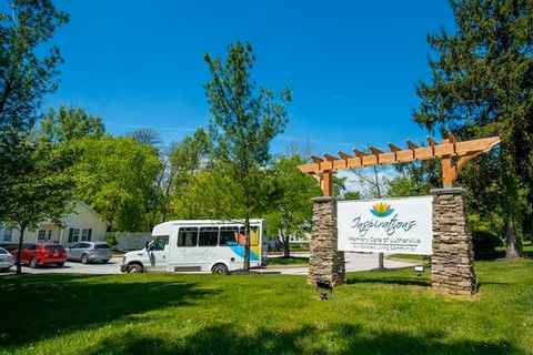 Outdoor view of Inspirations Memory Care of Lutherville facility sign mounted on stone pillars with a wooden pergola structure above. A white shuttle bus and several parked cars are visible in the background along with trees and a clear blue sky.