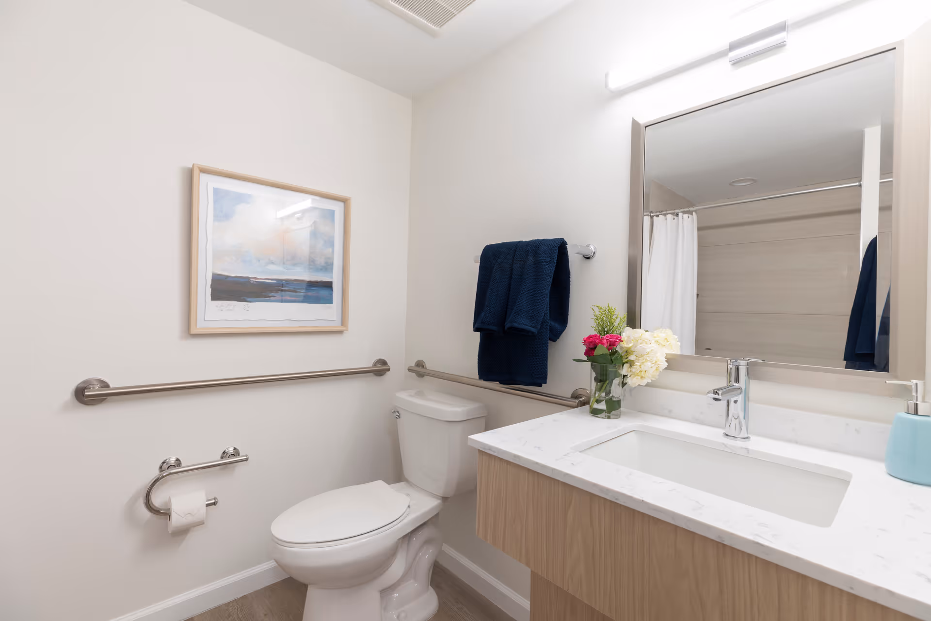 A clean and modern bathroom featuring a toilet with grab bars on the walls for accessibility, a white countertop with a built-in sink, a large mirror above the sink, a vase with flowers on the counter, a soap dispenser, and a framed artwork on the wall. There are dark blue towels hanging on a towel rack next to the toilet and a shower with a white curtain visible in the mirror reflection.