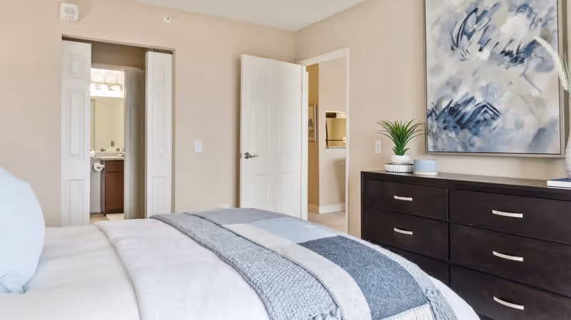 A neatly made bed with a blue and gray striped blanket in a bedroom. The room has beige walls, a dark wooden dresser with silver handles, a potted plant, and a large abstract painting above the dresser. Two open doors reveal a bathroom with a granite countertop and a hallway.