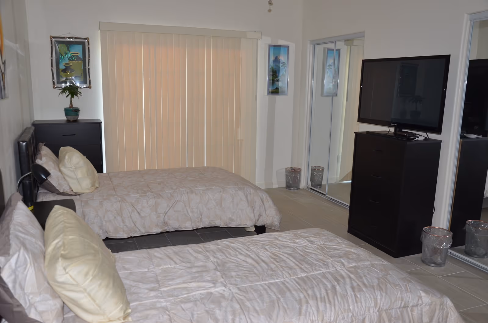 Two twin beds in a tidy bedroom with a TV on a dresser, vertical blinds and mirrored closet doors.