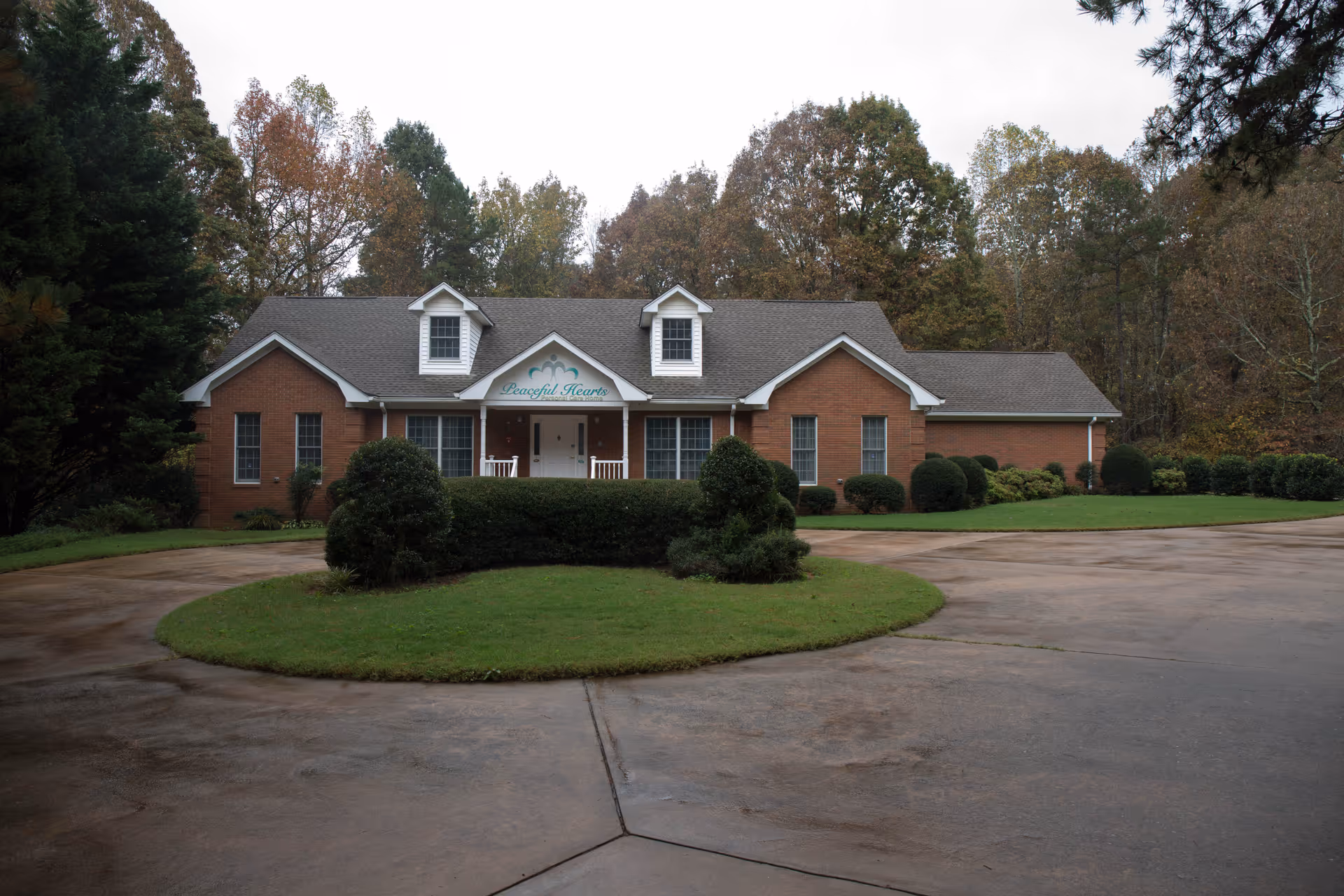 Brick single-story residential-style building with a front porch and circular driveway, landscaped shrubs and trees, with a sign reading "Peaceful Hearts" above the entrance.