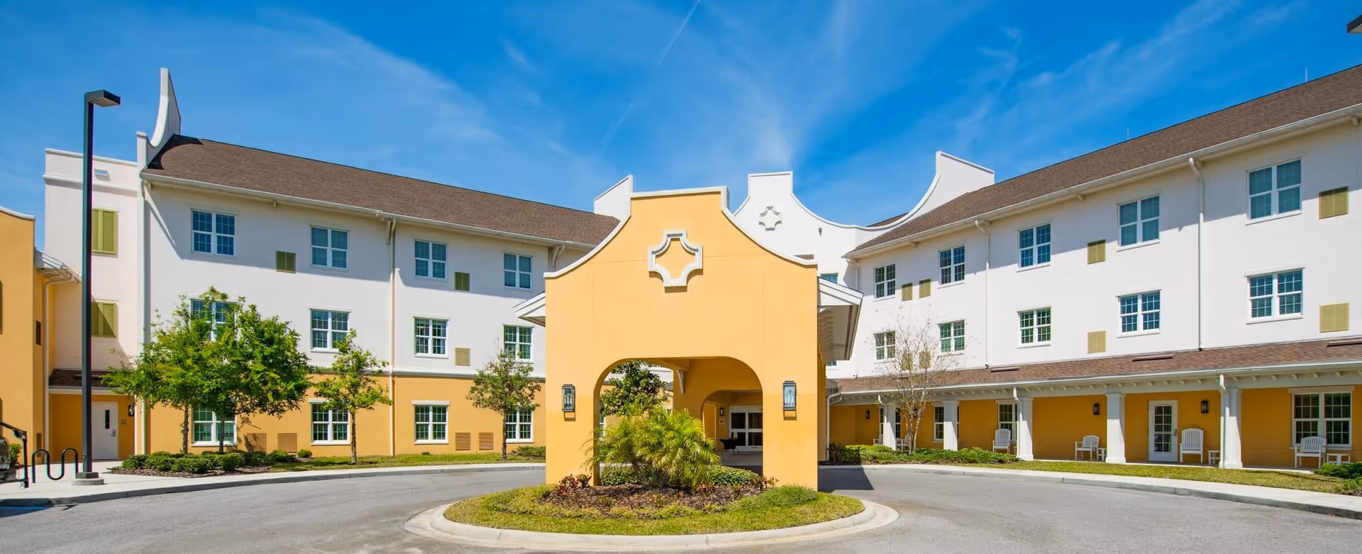 Exterior view of a senior living facility named Discovery Village Trinity, featuring a three-story building with white and yellow walls, multiple windows, and a covered entrance with decorative architectural details. There are small trees and landscaped greenery around the driveway under a clear blue sky.