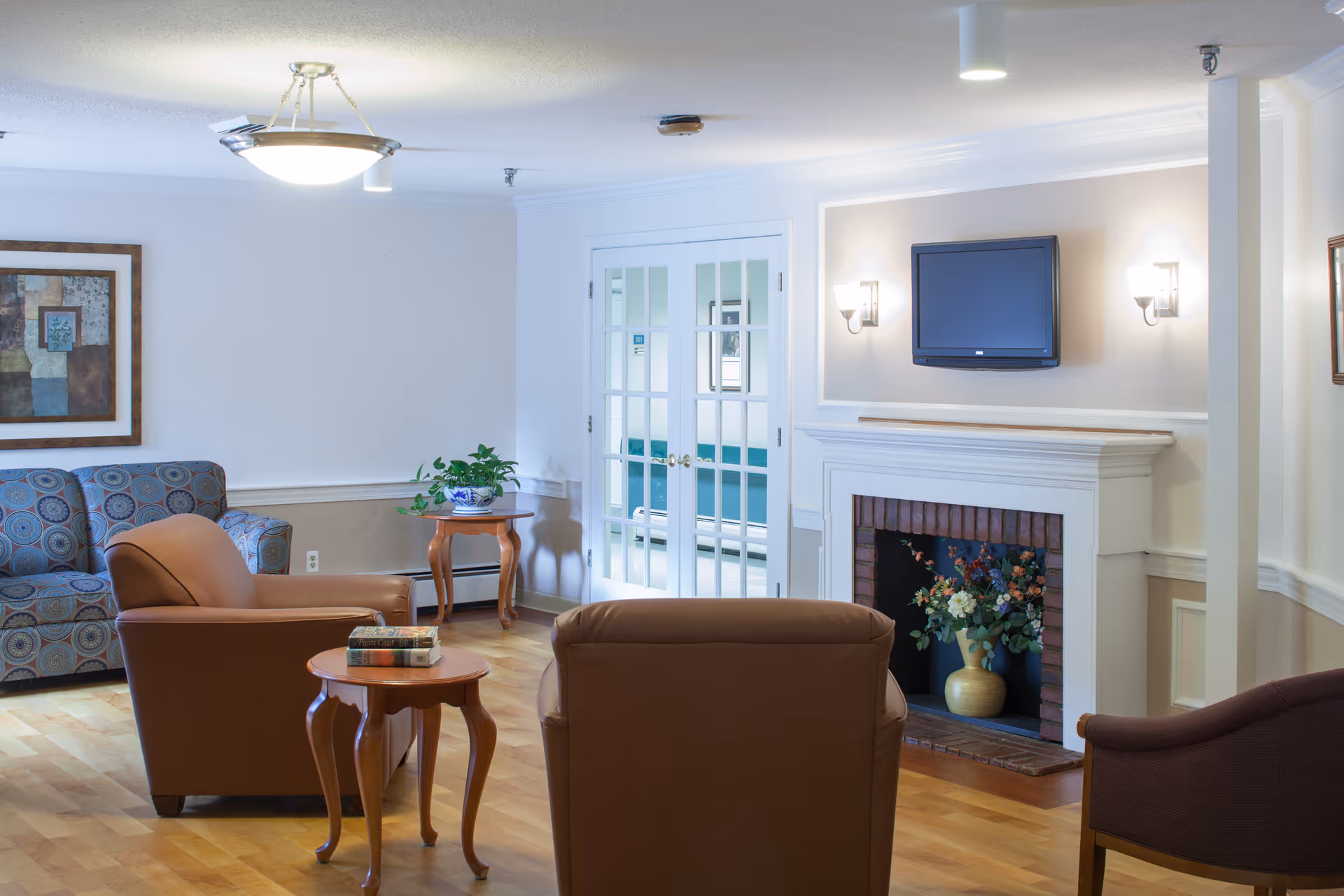A cozy living room area with wooden flooring, featuring a patterned sofa, two brown armchairs, a small wooden side table with books, and a wooden table with a potted plant. There is a white fireplace with a floral arrangement inside and a flat-screen TV mounted above it. French doors with glass panes lead to another room with teal seating.