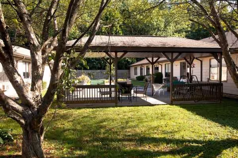 Outdoor courtyard area with a covered pavilion featuring tables and chairs, surrounded by single-story buildings and trees with green grass in the foreground.