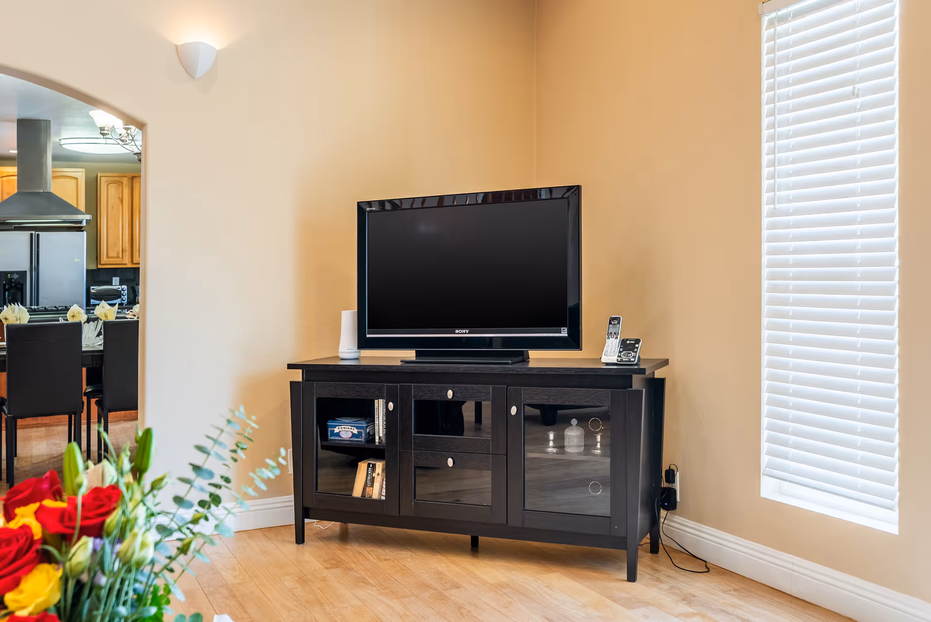 A living room corner featuring a black TV stand with a flat-screen Sony television on top. The stand has glass-paneled cabinets with books and decorative items inside. To the right is a window with white blinds, and to the left, an archway leads to a kitchen area with wooden cabinets and a dining table with black chairs. A bouquet of red and yellow flowers is partially visible in the foreground.