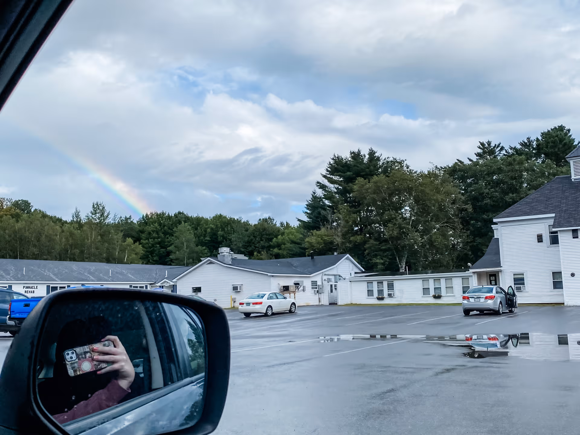 Exterior view of a white senior living building and parking lot with a rainbow in the sky and the photographer reflected in a car side mirror.